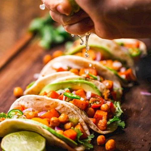 Tacos filled with chickpeas, vegetables, and garnished with lime being squeezed over them, placed on a wooden board.