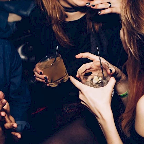 People are enjoying cocktails and laughing together, with close-up of hands and drinks at a social gathering, creating a lively atmosphere.