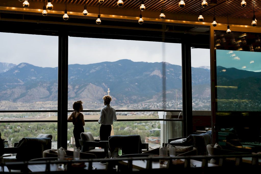 Two people stand by a restaurant/caf&eacute; window, looking out at mountain scenery as warm lights hang overhead.