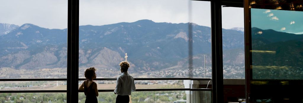 Two people stand by a restaurant/caf&eacute; window, looking out at mountain scenery as warm lights hang overhead.
