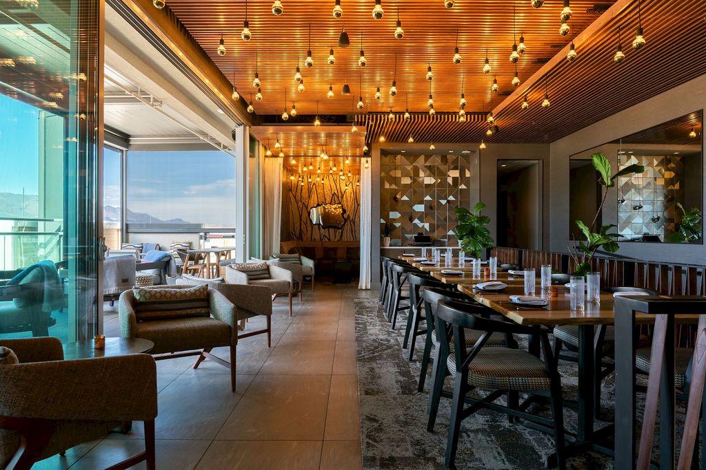 A modern restaurant lounge with warm wood ceiling, pendant lights, sofas, and a long dining table lined with plants.