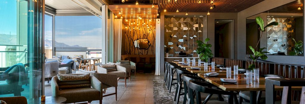 A modern restaurant lounge with warm wood ceiling, pendant lights, sofas, and a long dining table lined with plants.