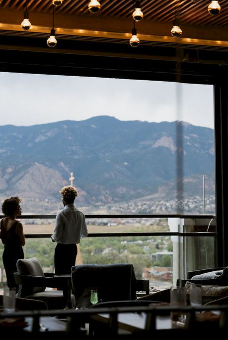 Two people sit in a caf&eacute; by large windows, looking at distant mountains under warm hanging lights.