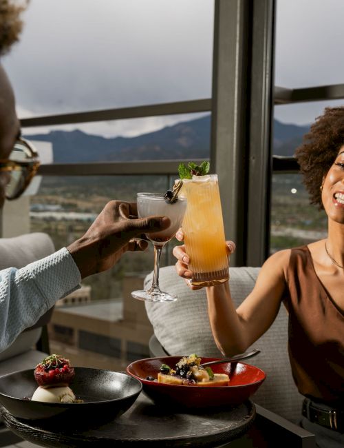 Two people clink cocktails on a patio restaurant table with appetizers, enjoying a mountain view outside the window.