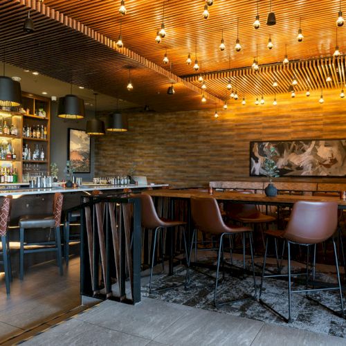 A modern bar interior with warm wood walls, hanging pendant lights, a liquor display, and high stools lined by the counter.