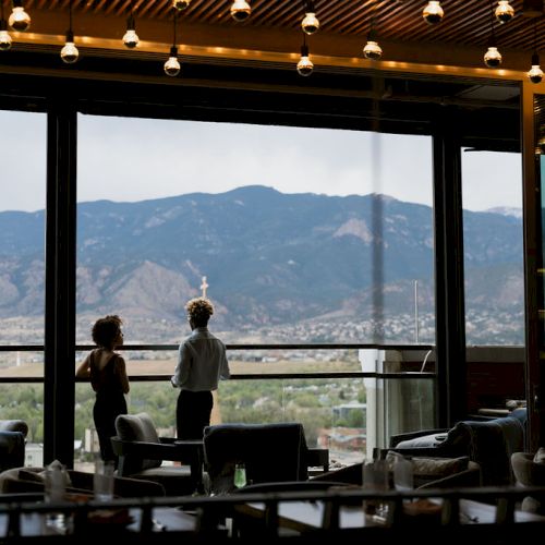 A couple sits in a cozy restaurant by large windows, watching mountains outside in the soft daylight.