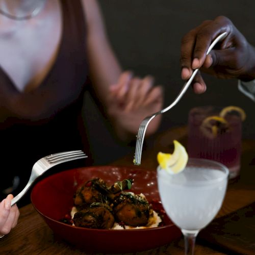 A diner forks saucy food from a red bowl while sharing the meal at a table with a drink nearby.