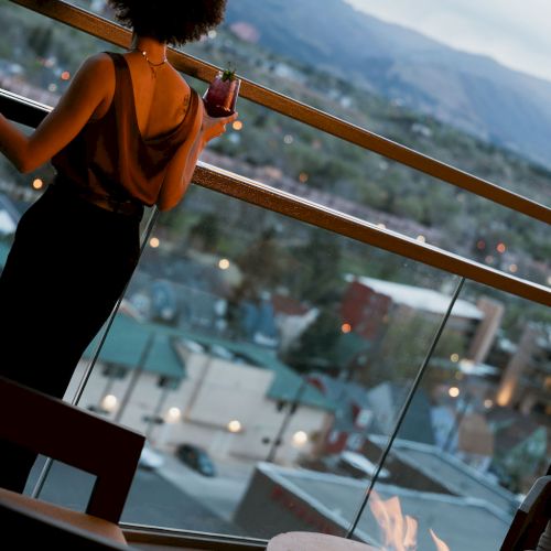A person on a balcony holds a cocktail with a drink near a lit fire, overlooking mountains and a valley below.