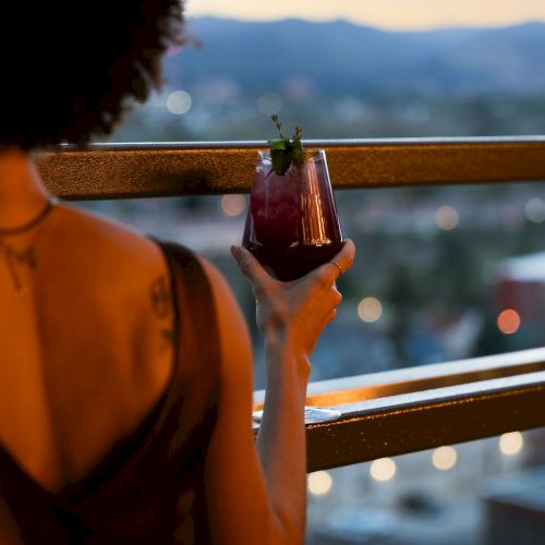 Someone holds a dark cocktail with mint on a terrace at sunset, gazing at mountains and city lights.