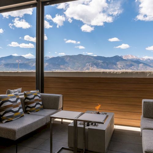 A hotel lounge with gray sofas by floor-to-ceiling windows, overlooking mountain ranges under a cloudy sky.