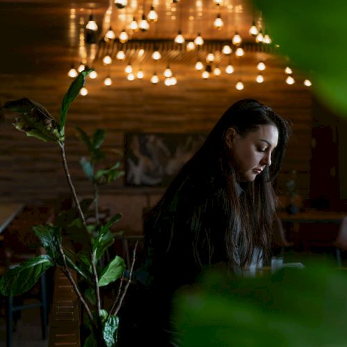 A woman sits in a cozy dim caf&eacute; with glowing string lights overhead, while a leafy plant in the foreground frames the scene.