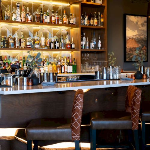 A cozy bar with shelves of liquor bottles, lit glassware, and dark leather booths with patterned chairs.