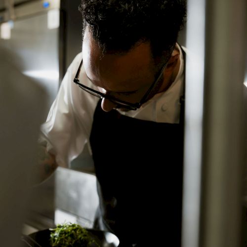 A chef in a dim kitchen leans over a bowl of food, plating and preparing a dish.
