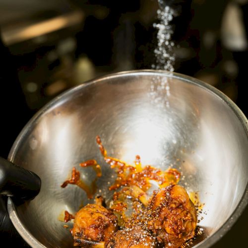 A black-gloved hand sprays sizzling sauce/oil into a stainless bowl, seasoning browned chicken pieces with spices and steam.