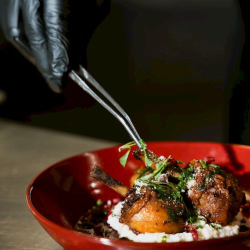 A chef in a dark apron plates savory lamb chops over creamy cheese and herbs in a red bowl.