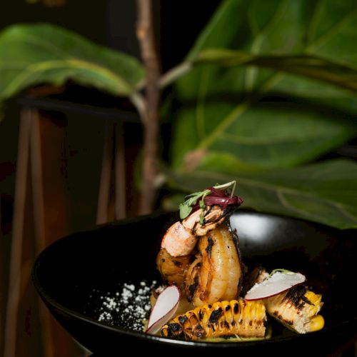 Grilled shrimp with roasted corn, radish slices, herbs, and spices served in a black bowl, lit with a dramatic green backdrop.