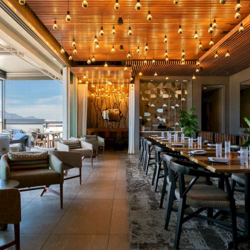 A modern restaurant/lounge with warm wooden ceiling, many hanging lights, upholstered seats, and long dining tables by big windows.