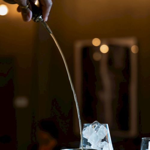 A bartender pours a clear drink into a metal cocktail cup with ice during a warm-lit bar service.