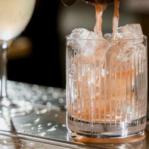 A bartender pours amber liquid over ice into a short glass on a bar tray.