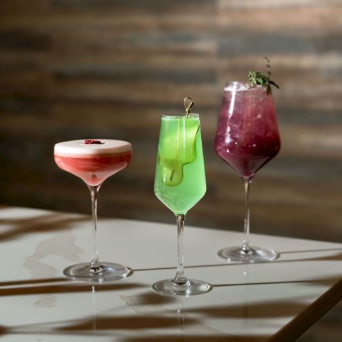 Three colorful cocktails in glasses&mdash;green, pink, and a frothy white-red&mdash;on a tabletop with a brick wall background.