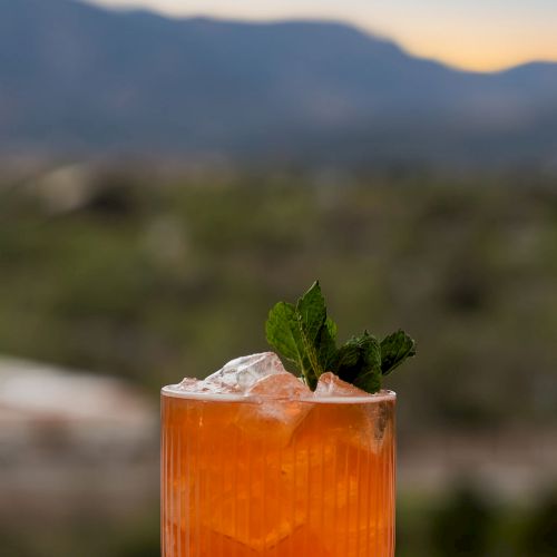 A tall orange cocktail with ice and fresh mint sits on a table, overlooking misty mountains at sunset.