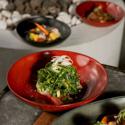 A red bowl of greens-topped salad sits in front of other plated dishes in a restaurant.