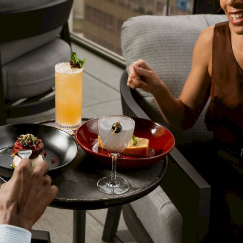 A couple cheers over a cocktail and shared small plates at a restaurant patio, smiling as dishes are served.