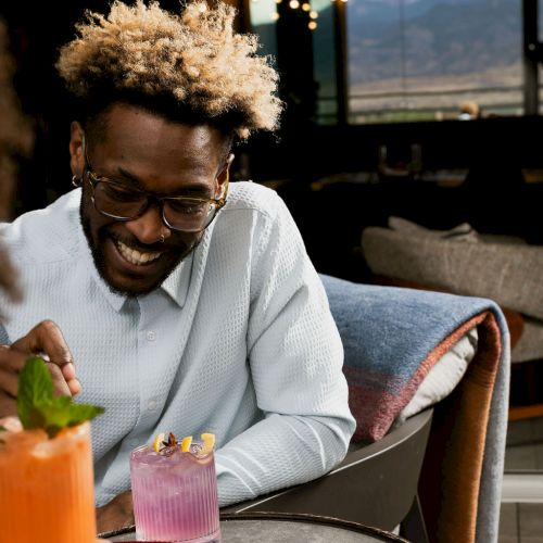 Smiling man with curly hair and glasses enjoys a meal at a caf&eacute; table, sipping from a pink cocktail garnished with fruit.