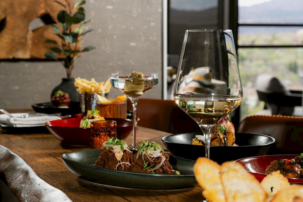 A dining table with plated food and two glasses of white wine, set indoors near a window with a scenic view outside.
