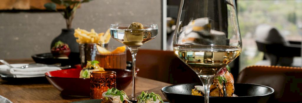 A dining table with plated food and two glasses of white wine, set indoors near a window with a scenic view outside.