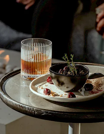 A cozy outdoor table with a lit drink, a small plant in a bowl, and a dessert on a plate near a glowing fire pit, stylish evening vibes.