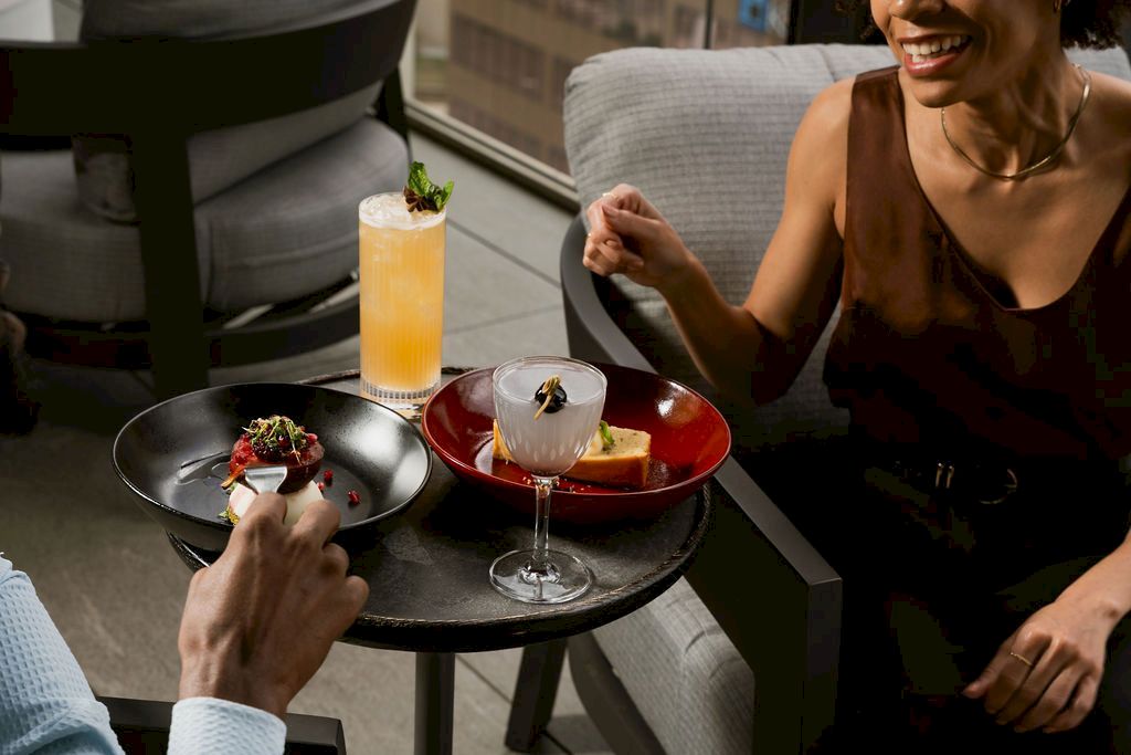 Two people dining on a patio, sharing cocktails and plates of food&mdash;one with an orange drink and garnished dish.