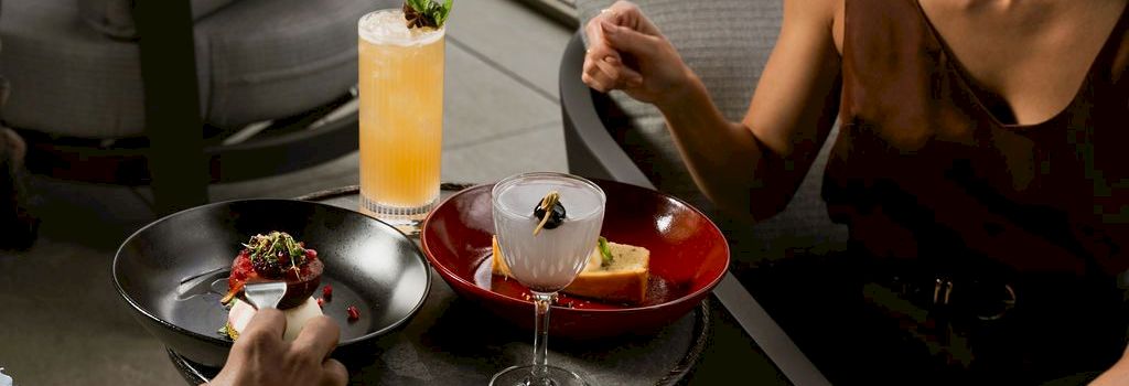 Two people dining on a patio, sharing cocktails and plates of food&mdash;one with an orange drink and garnished dish.