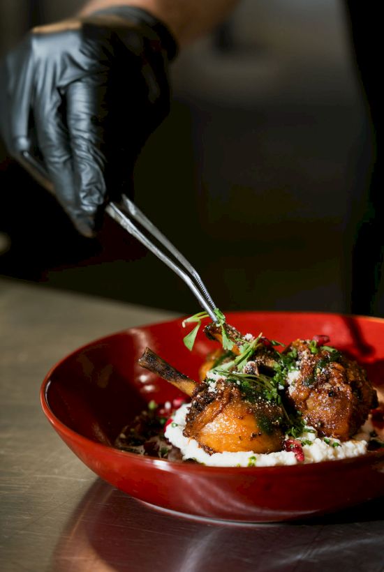 A chef in a black glove sprinkles herbs over plated lamb chops on a red bowl, with creamy sauce and greens.