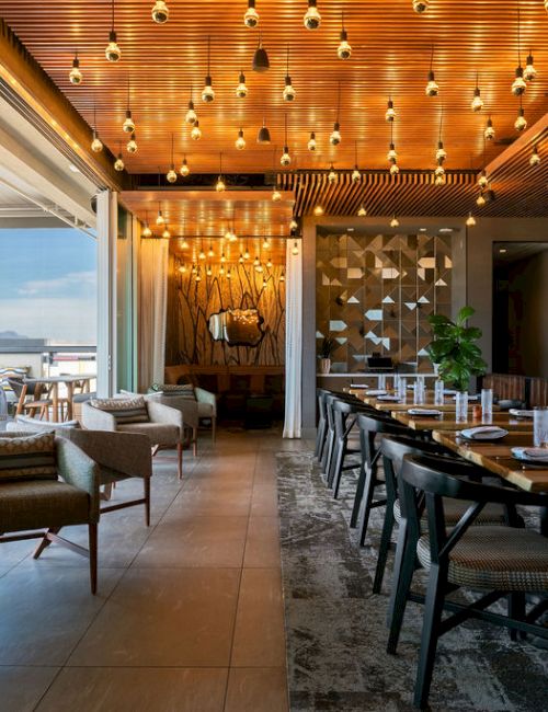 Modern restaurant lounge with warm hanging lights, cozy sofas, and a long dining table set with chairs.