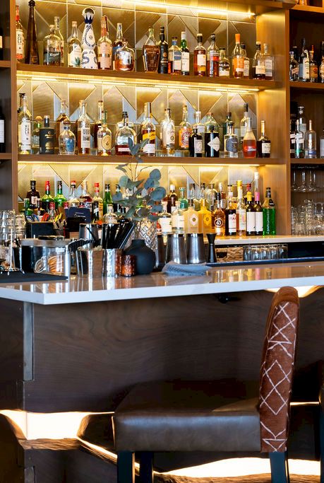 A stylish bar with shelves of liquor bottles, glowing backlighting, glassware, and leather stools arranged for drinks and cocktails.