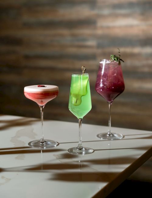 Three cocktails in stemmed glasses&mdash;green, red, and pale pink&mdash;garnished with fruit and herbs on a table.