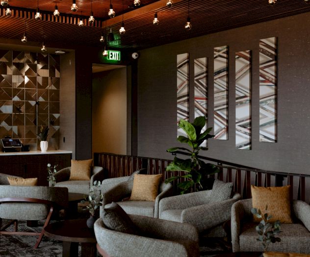 A modern lounge with rows of hanging globe lights, plush sofas, wall art panels, and a glowing EXIT sign above doors.