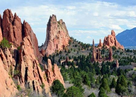 Towering red rock spires rise over a green valley under a bright blue sky, creating a dramatic desert mountain landscape.