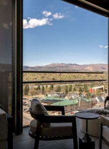 Cozy balcony seating by large windows, overlooking a town and mountain range under a blue sky.