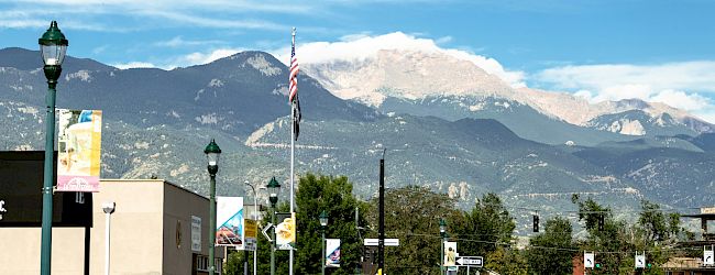 Mountain town street scene with cars, an American flag, green lampposts, and towering peaks under a bright blue sky.